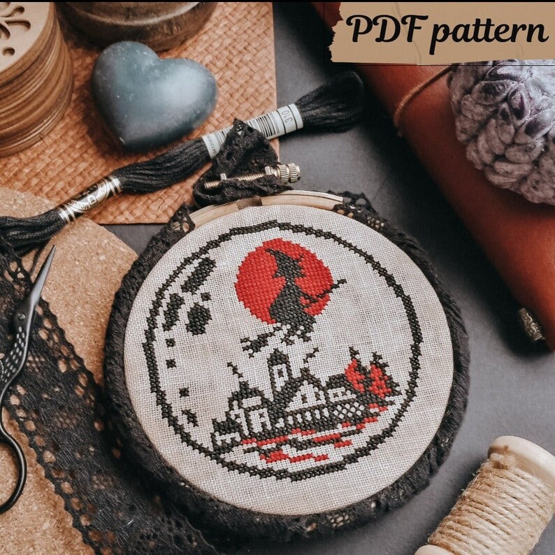 May include: A black and white cross-stitch pattern of a witch flying on a broomstick in front of a red moon and a silhouette of a house. The pattern is framed in a wooden hoop with black lace trim. The text "PDF pattern" is visible in the upper right corner of the image.