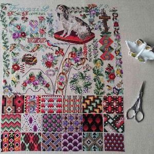 May include: A sampler embroidery on beige fabric featuring a central dog on a red cushion, surrounded by floral motifs, decorative borders, and geometric patterns. A white ceramic dish and silver metal scissors are placed to the right side.