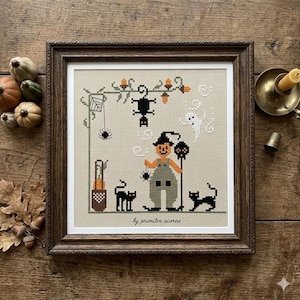 Może przedstawiać: Oprawiona sztuka haftu krzyżykowego z motywem Halloween. Grafika przedstawia stracha na wróble, czarne koty, pająki i ducha. Tekst "by primitive acorns" znajduje się na dole. Rama jest brązowa.
