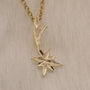 May include: A gold-colored necklace with a starburst pendant. The pendant has a star shape with multiple points and is suspended from a delicate chain. The necklace is displayed on a white, fluffy surface.