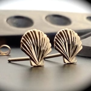 May include: A pair of gold-toned seashell stud earrings. The earrings are small and delicate, with a detailed design.