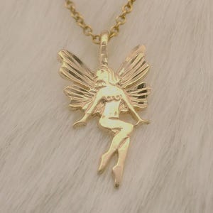 以下が含まれることがあります： 金色のペンダントネックレス。妖精のデザインで、翼を広げた妖精が繊細なチェーンから吊り下げられています。ネックレスは柔らかい白い背景に表示されています。