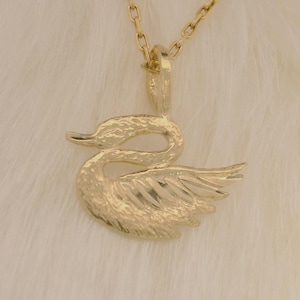 May include: A gold swan pendant on a necklace. The pendant is a detailed sculpture of a swan with its wings slightly spread. The necklace chain is gold, set against a white, fluffy background.