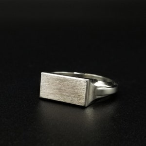 Peut inclure: Une bague signet en argent avec un dessus rectangulaire. Le dessus de la bague a une finition bross&eacute;e.