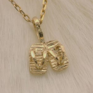 May include: A gold-colored necklace featuring a pendant shaped like a pair of flip-flops. The pendant and chain are made of a shiny, reflective metal. The necklace is displayed on a soft, white, textured surface.