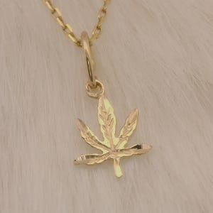 May include: A gold-colored pendant necklace featuring a cannabis leaf charm. The charm is a detailed, three-dimensional design of a cannabis leaf. The necklace chain is also gold-colored, and the background is a soft, white, textured surface.