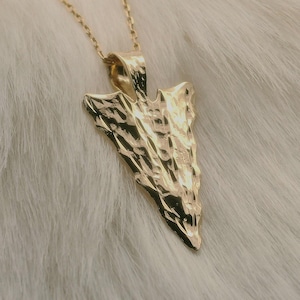 May include: A gold-colored pendant necklace featuring an arrowhead design. The pendant has a textured surface and is suspended from a delicate gold chain. The necklace is displayed on a white, fluffy surface.