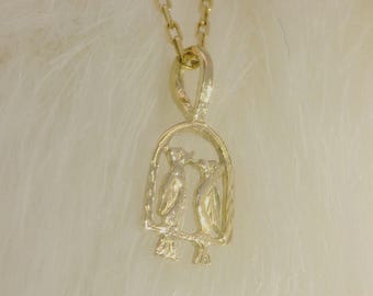 May include: A gold-colored pendant necklace featuring a detailed design of two birds perched on a branch within a decorative frame. The pendant hangs from a delicate chain, set against a soft, white background.