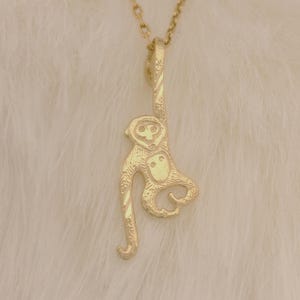 May include: A gold-colored monkey pendant necklace. The pendant is a detailed, stylized monkey design hanging from a delicate chain. The monkey has a long tail and is suspended against a soft, white background.