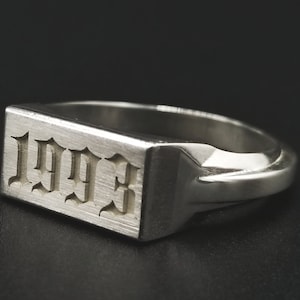 May include: A silver signet ring with a rectangular top featuring the year 1993 engraved in a gothic font.