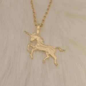 May include: A gold-colored unicorn pendant necklace. The pendant features a detailed unicorn design with a horn and flowing mane. The necklace is displayed on a white, fluffy surface.