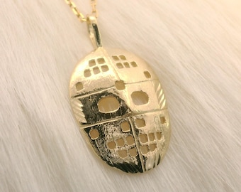 Könnte beinhalten: Goldfarbene Halskette mit Anhänger in Form einer Hockeymaske. Der Anhänger hat eine detaillierte, strukturierte Oberfläche mit Aussparungen. Die Halskette hängt an einer zarten Goldkette, vor weichem, weißem Hintergrund.
