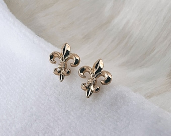 Könnte beinhalten: Ein Paar goldfarbene Fleur-de-lis-Ohrstecker. Die Ohrringe sind klein und zart und wären ein tolles Geschenk für jemanden, der die französische Kultur liebt.