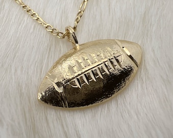 Könnte beinhalten: Eine goldene Kettenkette mit einem Fußballa Anhänger. Der Fußball ist mit Schnürsenkeln und einer strukturierten Oberfläche verziert.