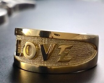 May include: A gold-colored ring with the word "LOVE" embossed on the front. The ring has a textured band and a polished finish. The background is dark, with a blurred silver object.