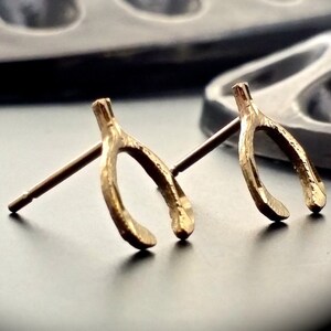 Puede incluir: Un par de pendientes de hueso de la suerte de color dorado. Los pendientes están hechos de metal texturizado y tienen un diseño simple y minimalista.