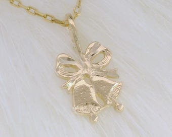 May include: A gold-coloured necklace featuring a pendant shaped like two bells with a bow. The chain and pendant are made of a shiny, metallic material. The necklace is displayed on a white, fluffy surface.