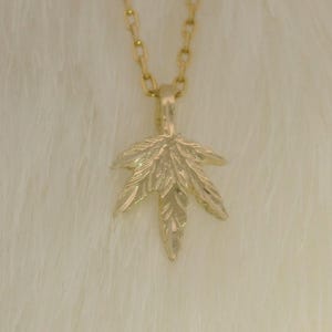 May include: A gold-colored necklace with a pendant shaped like a cannabis leaf. The leaf pendant is detailed with veining and hangs from a delicate chain. The necklace is displayed against a fluffy, white background.