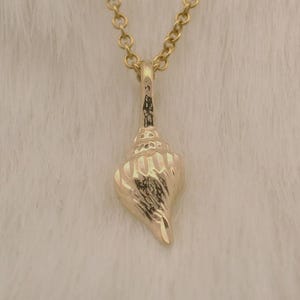 May include: A gold-colored necklace with a seashell pendant. The pendant is a detailed, textured seashell shape, suspended from a delicate chain. The necklace is set against a soft, white background.