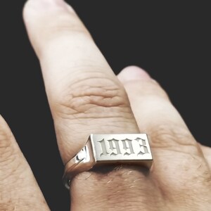 Peut inclure: Une bague en argent avec une bande rectangulaire grav&eacute;e de l'ann&eacute;e 1993 en police gothique.