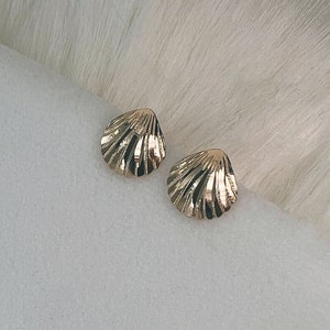 May include: A pair of gold-toned seashell-shaped earrings. The earrings are small and delicate, with a textured surface.