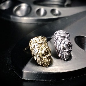 May include: Two lion head earrings, one gold and one silver, on a black surface.