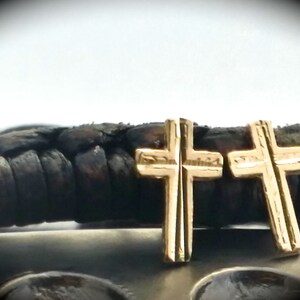 May include: Two gold-colored cross earrings. The crosses are detailed with vertical and horizontal lines. The earrings are displayed on a dark, textured surface, with a blurred background.
