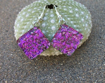 May include: A pair of iridescent purple glass earrings with silver hooks. The earrings are square shaped and have a textured surface.