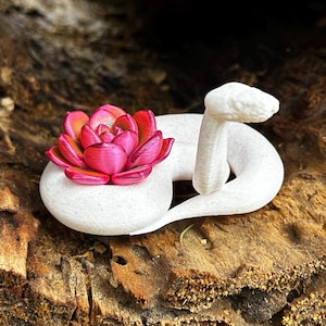 May include: A white, coiled snake figurine with a vibrant pink and red flower on its back. The snake is resting on a textured, brown wooden surface. The flower has multiple layers of petals, adding a touch of color to the scene.