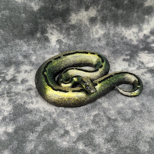 May include: A coiled snake figurine with a green and gold color scheme, accented with black stripes. The snake is posed on a gray, textured surface. The figurine has a detailed, realistic appearance.