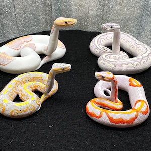 May include: Four decorative snake figurines in various colors, including white, orange, yellow, and gray. The snakes are coiled and have their heads raised, showcasing detailed patterns and textures. They are arranged on a black surface.