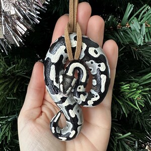 Peut inclure: Ornement de serpent en c&eacute;ramique peint &agrave; la main, avec un motif noir et blanc. L'ornement est suspendu par un cordon en cuir beige, parfait pour accrocher sur un sapin de No&euml;l ou comme &eacute;l&eacute;ment d&eacute;coratif. Le serpent est enroul&eacute; dans une pose d&eacute;corative.