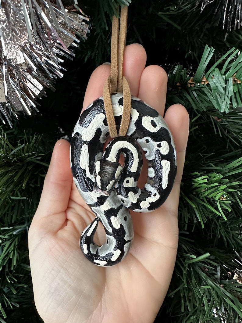Peut inclure: Ornement de serpent en c&eacute;ramique peint &agrave; la main, avec un motif noir et blanc. L'ornement est suspendu par un cordon en cuir beige, parfait pour accrocher sur un sapin de No&euml;l ou comme &eacute;l&eacute;ment d&eacute;coratif. Le serpent est enroul&eacute; dans une pose d&eacute;corative.