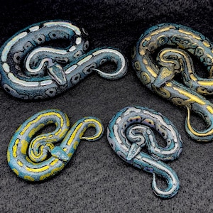May include: Four decorative snake figurines in various colors. The snakes are coiled and feature detailed patterns in shades of blue, yellow, and gold. The background is a dark, textured surface.