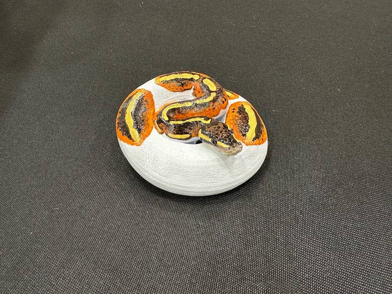 May include: A hand-painted, decorative stone shaped like a coiled snake. The snake has a brown and yellow pattern on a white base. The snake's head is visible, adding detail to the artwork. This item is a unique home decor piece.