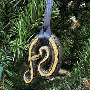 May include: A Christmas tree ornament featuring a coiled snake design. The ornament has a dark brown base with a yellow and black snake pattern. It hangs from a dark blue ribbon, set against a backdrop of green Christmas tree branches.