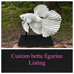 May include: A white, detailed figurine of a betta fish, mounted on a black stand. The fish has flowing fins and a textured surface. The text "Custom betta figurine Listing" is displayed below the figurine.