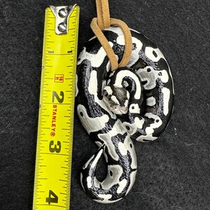 Peut inclure: Pendentif serpent enroul&eacute; avec un motif noir, blanc et gris, attach&eacute; &agrave; un cordon en cuir beige. Le pendentif mesure environ 7,6 cm de long. La t&ecirc;te du serpent est visible et le design est d&eacute;taill&eacute;. Le fond est gris fonc&eacute;.