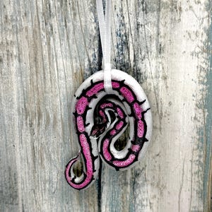 May include: A white, circular ornament with a pink and black snake design. The snake is coiled, with a detailed pattern. A white ribbon is attached for hanging. The ornament is set against a weathered, light-colored wood background.