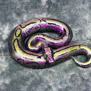 May include: A coiled, iridescent snake figurine with a blend of purple, green, and gold hues. The snake's body features a pattern of dark brown markings. The figurine is set against a textured gray background.