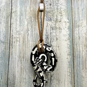 Peut inclure: Pendentif serpent en c&eacute;ramique avec un motif noir et blanc, suspendu par un cordon en cuir marron. Le pendentif est attach&eacute; &agrave; un petit carr&eacute; blanc. Le fond est une surface en bois bleu clair et gris.