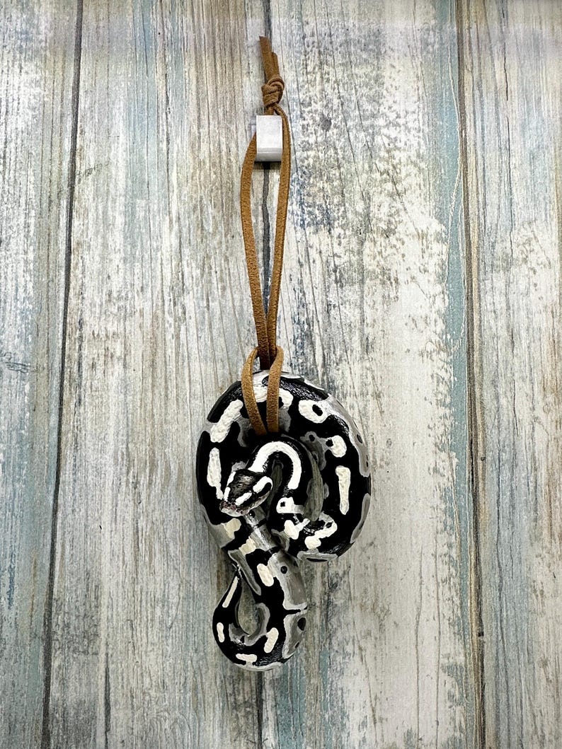 Peut inclure: Pendentif serpent en c&eacute;ramique avec un motif noir et blanc, suspendu par un cordon en cuir marron. Le pendentif est attach&eacute; &agrave; un petit carr&eacute; blanc. Le fond est une surface en bois bleu clair et gris.