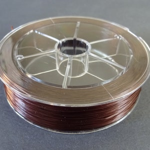 May include: A spool of dark brown fishing line. The line is tightly wound around a clear plastic spool. The spool has a clear, ridged design and a central hub. The fishing line appears to be a monofilament type.