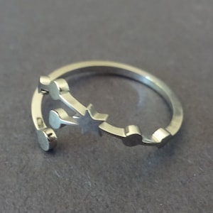 May include: A silver-colored ring with a star and small circular and rectangular accents. The ring has an open design, allowing for slight size adjustments. The ring is set against a gray background.