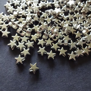 May include: A collection of small, silver-colored star-shaped charms scattered on a dark gray surface. The charms are metallic and have a five-pointed design, reflecting light. These decorative charms are suitable for various craft projects.