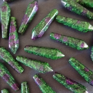 May include: A collection of green and pink gemstone points, also known as ruby zoisite, arranged on a dark surface. The points are faceted and have a natural, rough texture.