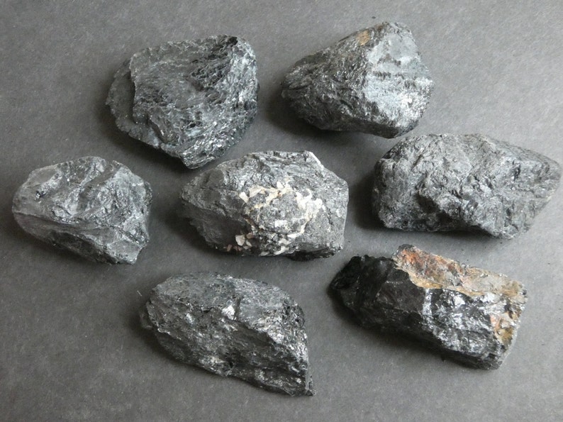 May include: Seven black, rough, and irregularly shaped stones. The stones have a slightly metallic sheen and are arranged on a gray surface.