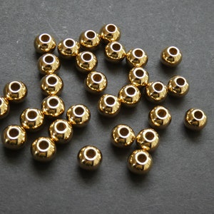May include: A collection of shiny, gold-colored round beads with a hole through the center. The beads are scattered on a dark gray surface, reflecting light. These beads are likely used for jewelry making or crafting.