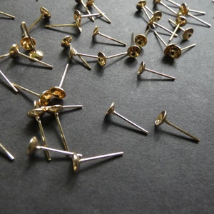 May include: A close-up of a group of gold-colored earring posts with round, flat tops. The posts are scattered on a black surface.