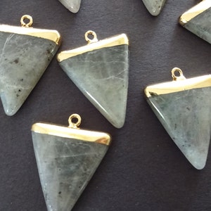 May include: A group of eight triangle-shaped pendants made of grey labradorite stone with gold-toned metal trim. Each pendant has a loop at the top for attaching to a chain or cord.